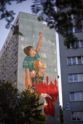 Zdjęcie pionowe przedstawiające boczną ścianę wielokondygnacyjnego budynku mieszkalnego, której powierzchnia została pokryta muralem z przedstawieniem figuratywnym. Na jego kompozycje składają się wiem postaci ludzkie – dziewczynki i chłopca w wieku nastoletnim, którzy zostali przedstawieni w sposób realistyczny. Siedzą oni na grzbiecie ogromnego koguta, którego grzebień przybrał formę gdańskich zabytków. Całość utrzymana jest raczej w ciemnej kolorystyce, której wyraźnym kontrapunktem jest krwistoczerwone korale i grzebień koguta.