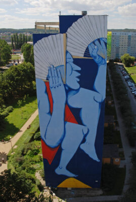 Zdjęcie pionowe przedstawiające boczną ścianę wielokondygnacyjnego budynku mieszkalnego, której powierzchnia została pokryta muralem z przedstawieniem ludzkim. Odrealniona, błękitna postać ukazana została w tanecznej pozie i trzyma ona w obu dłoniach dwa białe, azjatyckie wachlarze. Jeden z nich przysłania częściowo jej twarz. Cała kompozycja utrzymana jest w czystej i nasyconej kolorystyce. Zdominowana jest ona przez odcienie niebieskiego, zaś jej kontrapunktami są dwa geometryczne elementy: żółty oraz czerwony.