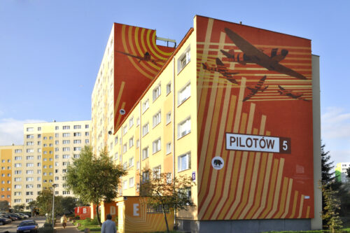 Zdjęcie poziome przedstawiające boczną ścianę wielokondygnacyjnego budynku mieszkalnego, której powierzchnia została pokryta muralem, którego estetyka przypomina sztukę komiksu. Przedstawia on wojskowe samoloty w czasie lotu. Zostały one przedstawione na abstrakcyjnym, geometrycznym tle, zaś kompozycja barwna utrzymana jest w nasyconej, intensywnej, pomarańczowo-żółtej kolorystyce.
