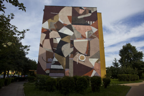 Zdjęcie poziome przedstawiające boczną ścianę wielokondygnacyjnego budynku mieszkalnego, której powierzchnia została pokryta muralem o abstrakcyjnym, geometrycznym charakterze. Cała kompozycja utrzymana jest w różnych odcieniach brązów. Całość przypomina abstrakcyjną mozaikę, na którą składają się różne figury geometryczne.