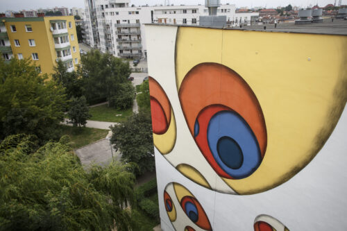 Zdjęcie poziome przedstawiające boczną ścianę wielokondygnacyjnego budynku mieszkalnego, której powierzchnia została pokryta muralem o abstrakcyjnym charakterze. Jego kompozycja zbudowana jest z trzech wielobarwnych okręgów rozmieszczonych równomiernie na powierzchni ściany. Całość utrzymana jest w ciepłej kolorystyce, zdominowanej przez nasycone czerwienie, żółcienie i pomarańcze, których kontrapunktami są pojedyncze błękitne i niebieskie okręgi.