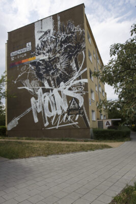 Zdjęcie pionowe przedstawiające boczną ścianę wielokondygnacyjnego budynku mieszkalnego, której powierzchnia została pokryta muralem o abstrakcyjnym charakterze. Jego kompozycja przypomina chaotyczny kolaż, na który składają się elementy roślinne, zwierzęce, a także pojedyncze napisy. Kolorystyka muralu utrzymana jest w różnych odcieniach brązów, która jest skontrastowana z białymi napisami i konturowaniem przedstawień figuratywnych.