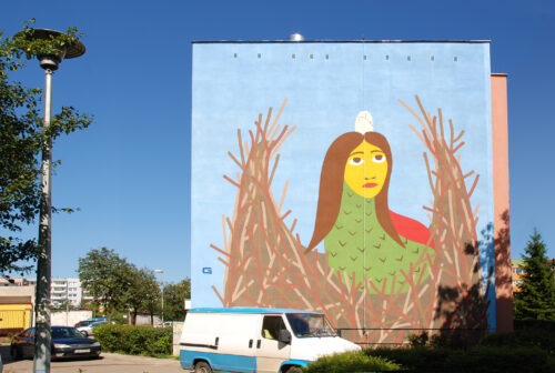 Zdjęcie poziome przedstawiające boczną ścianę wielokondygnacyjnego budynku mieszkalnego, której powierzchnia została pokryta kolorowym muralem. Przedstawia on hybrydę zwierzęco-ludzką – ptaka z głowa kobiety, która znajduje się w ptasim gnieździe. Mural utrzymany jest w jasnych (wręcz rozjaśnionych), czystych kolorach.