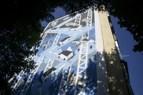 Zdjęcie poziome przedstawiające boczną ścianę wielokondygnacyjnego budynku mieszkalnego, której powierzchnia została pokryta muralem. Zostały na nim ukazane różnego rodzaju środki transportu: śmigłowce czy ciężarówki, a także astronauta czy satelita kosmiczny. Wszystkie elementy kompozycji są białe z wyraźnym, czarnym konturowaniem, przez co stanową kontrapunkty dla niebiesko-błękitnego tła. Całość utrzymana jest w komiksowej, a wręcz bajkowej stylistyce.