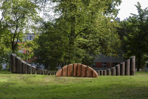 Zdjęcie poziome przedstawiające rzeźbę abstrakcyjną w przestrzeni parkowej. Rzeźba została wykonana w dwóch odcieniach brązowego drewna. Na jej konstrukcje składa się kilkanaście opracowanych pni drzew, które są ustawione w odpowiedniej kolejności zależnej od ich wysokości.