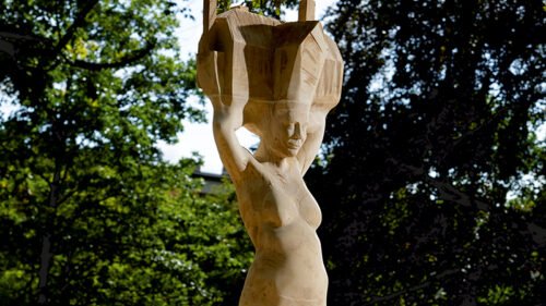 Zdjęcie pionowe przedstawiające nowoczesną rzeźbę w przestrzeni parkowej wykonaną w jasnobrązowym drewnie. Przedstawia ona realistycznie oddaną kobietę w bikini, która w uniesionych nad głową dłoniach trzyma konstrukcje domu jednorodzinnego. Postument rzeźby został wykonany w tym samym materiale i swoim wyglądem przypomina schematycznie przedstawiony dom składający się z czterech przestrzennych brył na planie rombu.