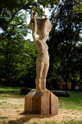 Zdjęcie pionowe przedstawiające nowoczesną rzeźbę w przestrzeni parkowej wykonaną w jasnobrązowym drewnie. Przedstawia ona realistycznie oddaną kobietę w bikini, która w uniesionych nad głową dłoniach trzyma konstrukcje domu jednorodzinnego. Postument rzeźby został wykonany w tym samym materiale i swoim wyglądem przypomina schematycznie przedstawiony dom składający się z czterech przestrzennych brył na planie rombu.