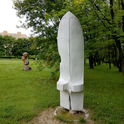 Zdjęcie przedstawiające rzeźbę abstrakcyjną w przestrzeni parkowej. Rzeźba została wykonana w drewnie, którego powierzchnia została pokryta białą farbą. Rzeźba sprawia wrażenie monolitycznej, jednorodnej a jej poszczególne elementy zostały zaznaczone jedynie poprzez zastosowanie mocniejszych rytów lub wypukłości.