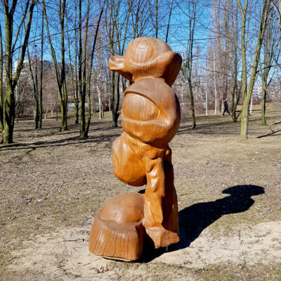Zdjęcie przedstawiające rzeźbę abstrakcyjną w przestrzeni parkowej. Rzeźba została wykonana w pomarańczowo-brązowym drewnie, którego powierzchnia została pozbawiona kory i wygładzona. Swoim wyglądem przypomina ona organiczne elementy, które wzajemnie się przenikają, tworzący tym samym pionową konstrukcję.