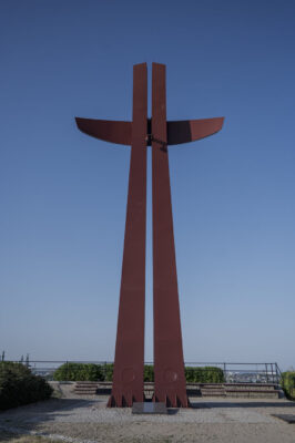 Zdjęcie pionowe przedstawiające wielkoformatową, metalową rzeźbę, która swoim kształtem przypomina krzyż rzymsko-katolicki, którego powierzchnia została pokryta ciemnobordową farbą. Rzeźba ma wydłużony kształt a jej krawędzie są subtelnie wygięte – przez co może sprawiać wrażenie lekkiej abstrakcyjności.