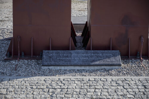 Zdjęcie poziome ze zbliżeniem na fragment metalowej rzeźby, której powierzchnia została pokryta ciemnobordową farbą. U jej podstawy została umieszczona ciemnoszara, marmurowa tablica z wyrytym napisem: „A gdy ja będę wywyższony nad ziemię, wszystkich do siebie przyciągnę. Jan 8, 28”.