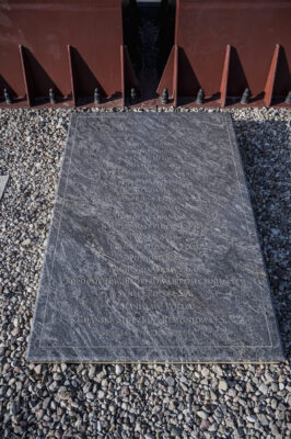 Zdjęcie pionowe ze zbliżeniem na fragment metalowej rzeźby, której powierzchnia została pokryta ciemnobordową farbą. U jej podstawy została umieszczona ciemnoszara, marmurowa tablica upamiętniająca fundatorów pomnika „Krzyż Millenijny – pomnik 2000-lecia chrześcijaństwa i 1000-lecia Gdańska”.