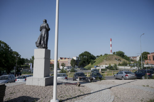 Zdjęcie pionowe przedstawiające rzeźbę w przestrzeni miejskiej. Figura mężczyzny została oddana w sposób realistyczny, a draperie sutanny, w którą jest on ubrany układają się w zgodny sposób z anatomią. Rzeźba została umieszczona na wysokim, prostopadłościennym postumencie.