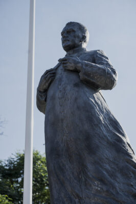 Zdjęcie pionowe przedstawiające zbliżenie na rzeźbę w przestrzeni miejskiej. Figura mężczyzny ubranego w sutannę, została oddana w sposób realistyczny. Postać stoi wyprostowana i patrzy pewnie przed siebie. W swojej prawej dłoni trzyma krzyż na wysokości klatki piersiowej, zaś wskazującym palcem lewej dłoni na niego wskazuje.