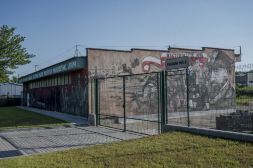 Zdjęcie poziome przedstawiające jednopiętrowy, niski budynek w przestrzeni industrialnej, którego jedna ze ścian bocznych została pokryta muralem o tematyce historycznej. Przedstawia on walczących żołnierzy w czasie walk o Westerplatte w 1939 r. Scena została oddana w sposób realistyczny a kompozycja jest zdominowana przez różne odcienie beżu i brązu – przez co przypomina ona kolorystycznie zdjęcie wykonane techniką sepii. Pojawia się także element biało-czerwony, czyli polska flaga, na której znajduje się napis: „Westerplatte 1939”.