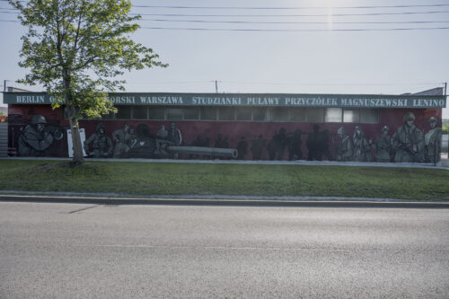 Zdjęcie poziome przedstawiające jednopiętrowy, niski budynek w przestrzeni industrialnej, którego jedna ze ścian bocznych została pokryta muralem o tematyce historycznej. Przedstawia on polskich żołnierzy na bordowym tle. Postaci zostały oddane w sposób realistyczny, podobnie jak umundurowanie i uzbrojenie. W górnej części kompozycji zostały wypisane nazwy miejscowości niektórych walk w czasie II Wojny Światowej, jak np. Lenino, Warszawa czy Puławy.