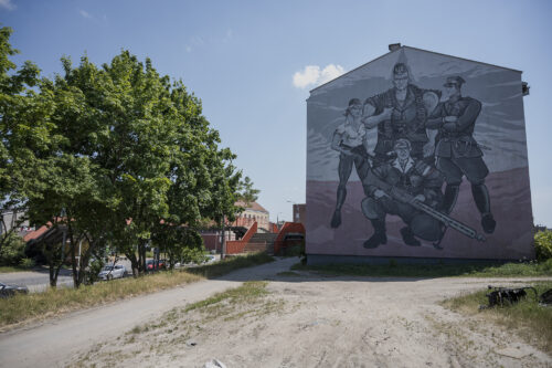 Zdjęcie poziome przedstawiające ścianę boczną jednego z zabytkowych budynków w przestrzeni miejskiej. Jest ona w całości pokryta wielkoformatowym muralem z przedstawieniami ludzkimi. Poprzez nadanie postaciom bardzo wydłużonych wręcz odrealnionych proporcji ciała a także silnie rozbudowanej muskulatury, mural przypomina swoją stylistyką dzieła komiksowe. Przedstawia on czterech żołnierzy nie tle polskiej flagi.