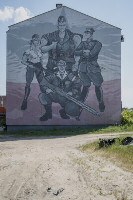 Zdjęcie pionowe przedstawiające ścianę boczną jednego z zabytkowych budynków w przestrzeni miejskiej. Jest ona w całości pokryta wielkoformatowym muralem z przedstawieniami ludzkimi. Poprzez nadanie postaciom bardzo wydłużonych wręcz odrealnionych proporcji ciała a także silnie rozbudowanej muskulatury, mural przypomina swoją stylistyką dzieła komiksowe. Przedstawia on czterech żołnierzy nie tle polskiej flagi.