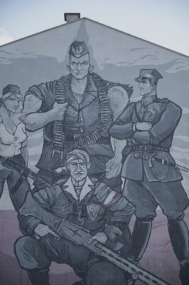 Zdjęcie pionowe ze zbliżeniem na fragment muralu pokrywającym jedną ze ścian bocznych zabytkowego budynku w przestrzeni miejskiej. Jest ona w całości pokryta wielkoformatowym muralem z przedstawieniami ludzkimi. Poprzez nadanie postaciom bardzo wydłużonych wręcz odrealnionych proporcji ciała a także silnie rozbudowanej muskulatury, mural przypomina swoją stylistyką dzieła komiksowe. Przedstawia on czterech żołnierzy nie tle polskiej flagi.