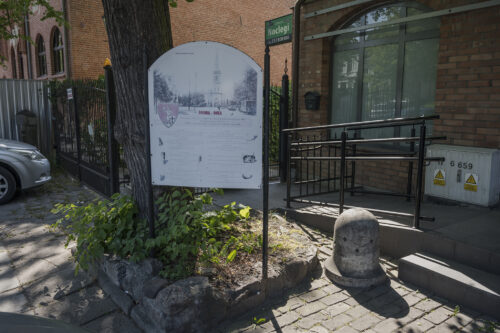 Zdjęcie poziome ze zbliżeniem na tablice informacyjną zwieńczoną łukowo. Jest ona ustawiona na wysokich prętach pod wieloletnim drzewem.