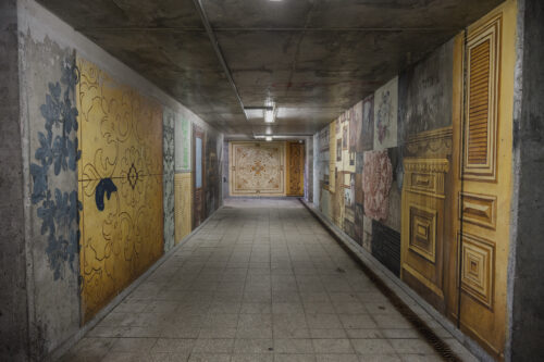 Zdjęcie poziome przedstawiające betonowe przejście podziemne, którego ściany pokryte są wieloma muralami, które nawiązują swoją formą do różnych ozdobnych płyt podłogowych a także elementów architektonicznych. Cała kompozycja utrzymana jest w różnych odcieniach żółcieni i pomarańczy, jednak pojawiają się również elementy w różu lub bieli. Całość przypomina bogatą i bardzo wzorzystą mozaikę.