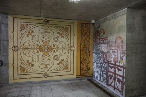 Zdjęcie poziome przedstawiające zbliżenie na róg ścian betonowego przejścia podziemnego. Ściany pokryte są wieloma muralami, które nawiązują swoją formą do różnych ozdobnych płyt podłogowych a także elementów architektonicznych. Cała kompozycja utrzymana jest w różnych odcieniach żółcieni i pomarańczy, jednak pojawiają się również elementy w różu lub bieli. Całość przypomina bogatą i bardzo wzorzystą mozaikę.