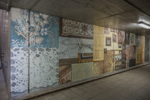 Zdjęcie poziome przedstawiające zbliżenie jedną ze ścian betonowego przejścia podziemnego. Pokryta jest ona wieloma muralami, które nawiązują swoją formą do różnych ozdobnych płyt podłogowych a także elementów architektonicznych. Cała kompozycja utrzymana jest w różnych odcieniach żółcieni i pomarańczy, jednak pojawiają się również elementy w różu, bieli czy turkusu. Całość przypomina bogatą i bardzo wzorzystą mozaikę.