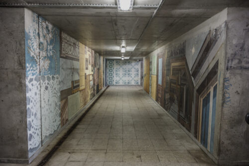 Zdjęcie poziome przedstawiające betonowe przejście podziemne, którego ściany pokryte są wieloma muralami, które nawiązują swoją formą do różnych ozdobnych płyt podłogowych a także elementów architektonicznych. Cała kompozycja utrzymana jest w różnych odcieniach żółcieni i pomarańczy, jednak pojawiają się również elementy w różu, bieli czy turkusu. Całość przypomina bogatą i bardzo wzorzystą mozaikę.