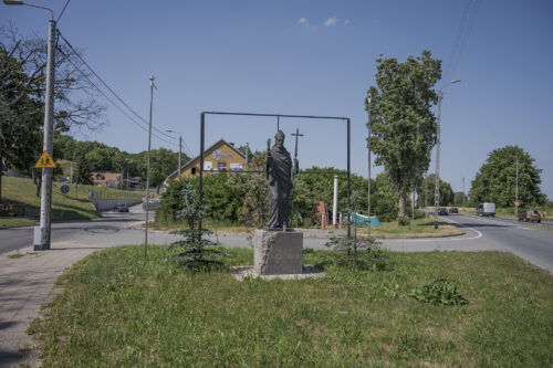 Zdjęcie poziome przedstawiające rzeźbę kapłana ustawioną na sześciennym postumencie. Pomnik znajduje się na środku skrzyżowania ulic. Postać kapłana stoi wyprostowana i patrzy pewnie przed siebie. W swojej lewej dłoni trzyma długi pastorał, natomiast prawą dłoń ma wysoko uniesioną w geście przywitania. Na jednej ze ścian postumentu został umieszczony napis: „Święty Wojciech 997”.