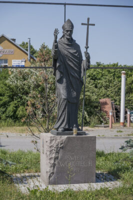 Zdjęcie pionowe przedstawiające rzeźbę kapłana ustawioną na sześciennym postumencie. Pomnik znajduje się na trawniku niedaleko ulicy. Postać kapłana stoi wyprostowana i patrzy pewnie przed siebie. W swojej lewej dłoni trzyma długi pastorał, natomiast prawą dłoń ma wysoko uniesioną w geście przywitania. Na jednej ze ścian postumentu został umieszczony napis: „Święty Wojciech 997”. Figura została wykonana w brązie, natomiast postument w jasnoróżowym granicie.
