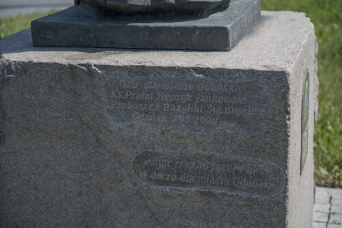 Zdjęcie poziome ze zbliżeniem na fragment sześciennego postumentu rzeźby, który został wykonany w jasnoróżowym granicie. Na jednej z jego ścian został wyryty napis upamiętniający podarowanie rzeźby przez ks. Jankowskiego dla miasta Gdańska.