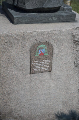 Zdjęcie poziome ze zbliżeniem na fragment sześciennego postumentu rzeźby, który został wykonany w jasnoróżowym granicie. Na jednej z jego ścian została umieszczona tabliczka o łukowym zakończeniu, na której znajduje się napis upamiętniający księdza Głodzia oraz kolorowy herb arcybiskupi.