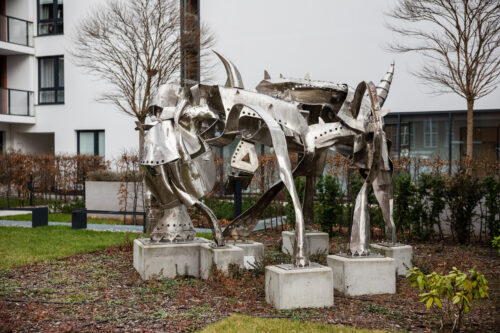 Zdjęcie przedstawiające przestrzenną instalację artystyczną, która została wykonana ze stali. Jej konstrukcja przypomina swoim kształtem wielonożne zwierzę o odrealnionej, abstrakcyjnej formie. Figura usytułowana jest na pięciu, niskich, betonowych postumentach. Wykorzystana stal przypomina swoim wyglądem rycerską, średniowieczną zbroję.