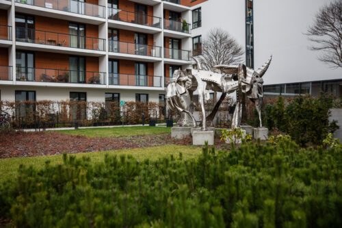 Zdjęcie przedstawiające przestrzenną instalację artystyczną, która została wykonana ze stali. Jej konstrukcja przypomina swoim kształtem wielonożne zwierzę o odrealnionej, abstrakcyjnej formie. Figura usytułowana jest na pięciu, niskich, betonowych postumentach. Wykorzystana stal przypomina swoim wyglądem rycerską, średniowieczną zbroję.