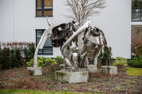 Zdjęcie przedstawiające przestrzenną instalację artystyczną, która została wykonana ze stali. Jej konstrukcja przypomina swoim kształtem wielonożne zwierzę o odrealnionej, abstrakcyjnej formie. Figura usytułowana jest na pięciu, niskich, betonowych postumentach. Wykorzystana stal przypomina swoim wyglądem rycerską, średniowieczną zbroję.