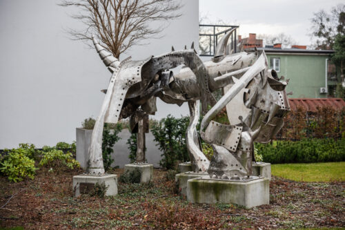 Zdjęcie przedstawiające przestrzenną instalację artystyczną, która została wykonana ze stali. Jej konstrukcja przypomina swoim kształtem wielonożne zwierzę o odrealnionej, abstrakcyjnej formie. Figura usytułowana jest na pięciu, niskich, betonowych postumentach. Wykorzystana stal przypomina swoim wyglądem rycerską, średniowieczną zbroję.
