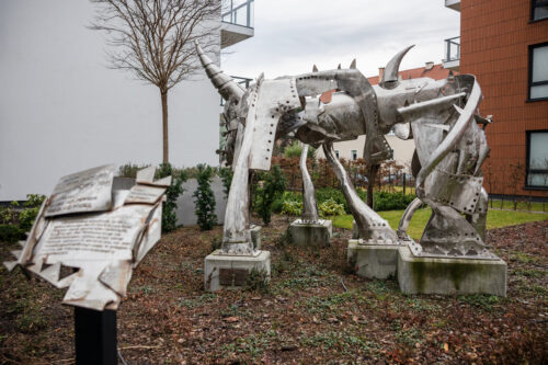 Zdjęcie przedstawiające przestrzenną instalację artystyczną, która została wykonana ze stali. Jej konstrukcja przypomina swoim kształtem wielonożne zwierzę o odrealnionej, abstrakcyjnej formie. Figura usytułowana jest na pięciu, niskich, betonowych postumentach. Wykorzystana stal przypomina swoim wyglądem rycerską, średniowieczną zbroję.