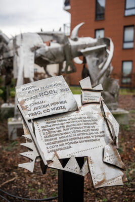 Zdjęcie przedstawiające zbliżenie na przestrzenna konstrukcję, na którą składają się stalowe tabliczki z wyrytymi napisami w prostej, technicznej czcionce. Nachodzą one na siebie wzajemnie, tworząc tym samym efekt chaotyczności. Na jednej z tabliczek znajduje się informacja o autorze pracy, jej tytuł oraz nazwa cyklu: Elvin Flamingo, Robot Roel, 2021, z cyklu Jestem pewien tego co widzę.