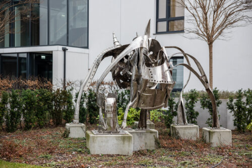 Zdjęcie przedstawiające przestrzenną instalację artystyczną, która została wykonana ze stali. Jej konstrukcja przypomina swoim kształtem wielonożne zwierzę o odrealnionej, abstrakcyjnej formie. Figura usytułowana jest na pięciu, niskich, betonowych postumentach. Wykorzystana stal przypomina swoim wyglądem rycerską, średniowieczną zbroję.