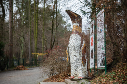 Na fotografii znajduje się rzeźba przedstawiająca pingwina w tle widoczna jest tablica informująca o tym, że jest on na terenie Gdańskiego Ogrodu Zoologicznego. Ptak zwrócony jest dziobem w prawą stronę kadru i znajduje się w jego centrum. Powierzchnię rzeźby wypełnia mozaika składająca się z ceramicznych kostek w kolorach brązu, żółci, czerni i bieli. Zarysowują one oczy, dziób i pozostałe elementy upierzenia pingwina. Widoczny jest mały postument na którym znajduje się rzeźba.