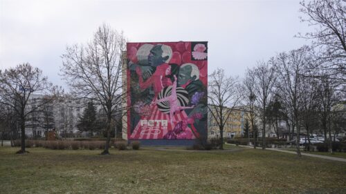 Zdjęcie poziome przedstawiające boczną ścianę bloku mieszkalnego, która w całości została pokryta kolorowym muralem. Malowidło ścienne upamiętnia 25 rocznicę festiwalu FETA, czyli Międzynarodowego Festiwalu Teatrów Plenerowych i Ulicznych FETA. Mural został wykonany w komiksowej stylistyce i utrzymany jest w jasnoróżowej i kontrastowej, ciemnozielonej kolorystyce.