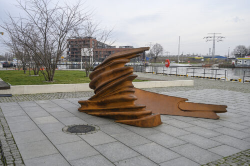 Zdjęcie poziome ukazujące pod kątem, niewielkich rozmiarów, abstrakcyjną rzeźbę w przestrzeni miejskiej. Swoim wyglądem przypomina ona zgniecioną, zardzewiałą stal, która układa się łukowo niczym fala morska. W tle widoczna jest zabudowa miejska a także kanał wodny.