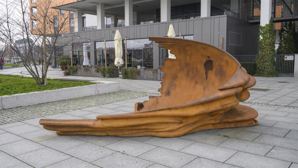 Zdjęcie poziome ukazujące pod kątem, niewielkich rozmiarów, abstrakcyjną rzeźbę w przestrzeni miejskiej. Swoim wyglądem przypomina ona zgniecioną, zardzewiałą stal, która układa się łukowo niczym fala morska. W tle widoczna jest zabudowa miejska.