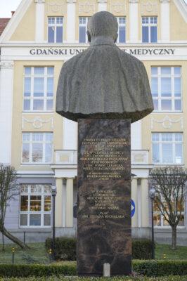 Zdjęcie pionowe, na którym znajduje się popiersie ustawione na wysokim, prostopadłościennym, marmurowym postumencie. Rzeźba przedstawia mężczyznę w średnim wieku. Na ścianie postumentu wyryte są złote litery, które układając się w m.in. napis: W 50 rocznicę utworzenia Akademii Medycznej w Gdańsku. Skwer ten otrzymał nazwę „Plac dr Stefana Michalaka”. W tle widoczny jest zabytkowy budynek Akademii Medycznej w Gdańsku.