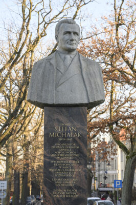 Zdjęcie pionowe, na którym znajduje się popiersie ustawione na prostopadłościennym, marmurowym postumencie. Rzeźba przedstawia uśmiechniętego mężczyznę w średnim wieku ubranego w garnitur. Rzeźba ustawiona jest na wysokim, prostopadłościennym, marmurowym postumencie. Na jednej z jego ścian wyryte są złote litery, które układając się m.in. w napis: Dr med. Stefan Michalak , 1909-1947. Organizator i pierwszy dyrektor szpitala, współtwórca Akademii Medycznej w Gdańsku.