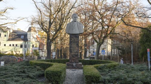 Zdjęcie poziome, na którym znajduje się popiersie ustawione na prostopadłościennym, marmurowym postumencie. Rzeźba przedstawia uśmiechniętego mężczyznę w średnim wieku ubranego w garnitur. Rzeźba ustawiona jest na wysokim, prostopadłościennym, marmurowym postumencie. Na jednej z jego ścian wyryte są złote litery. W tle widoczna jest zabytkowa zabudowa Gdańska.