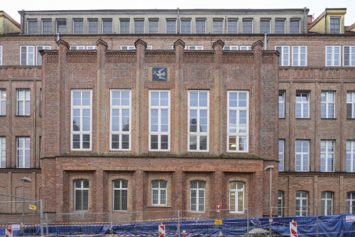 Zdjęcie poziome, wykonane na wprost przedstawiające modernistyczny budynek z czerwonej cegły. Nad jednym z pięciu długich, wąskich okien została umiejscowiona mozaika z alegorycznym przedstawieniem gołąbka pokoju z gałązką oliwną w dziobie.