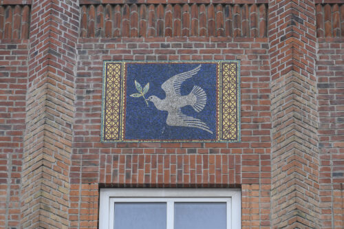 Zdjęcie poziome ze zbliżeniem na mozaikę z alegorycznym przedstawieniem gołąbka pokoju z gałązką oliwną w dziobie. Znajduje się on na ciemnoniebieskim tle, które jest po obydwu stronach ozdobione złotą dekoracją geometryczną. Mozaika jest wmontowana w ceglaną, czerwoną ścianę budynku.
