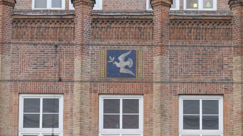 Zdjęcie poziome ze zbliżeniem na mozaikę z alegorycznym przedstawieniem gołąbka pokoju z gałązką oliwną w dziobie. Znajduje się on na ciemnoniebieskim tle, które jest po obydwu stronach ozdobione złotą dekoracją geometryczną. Mozaika jest wmontowana w ceglaną, czerwoną ścianę budynku.