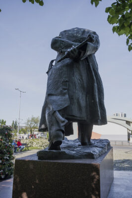 Zdjęcie wykonane na wprost, przedstawiające całopostaciową, rzeźbę mężczyzny w podeszłym wieku. Pomnik ustawiony jest na niskim, prostopadłościennym postumencie wykonanym z czerwonego granitu. Figura mężczyzny ubrana jest w mundur oraz długi płaszcz. Mężczyzna jest lekko pochylony przed siebie, natomiast dłonie ma splecione za plecami, w których trzyma buławę.